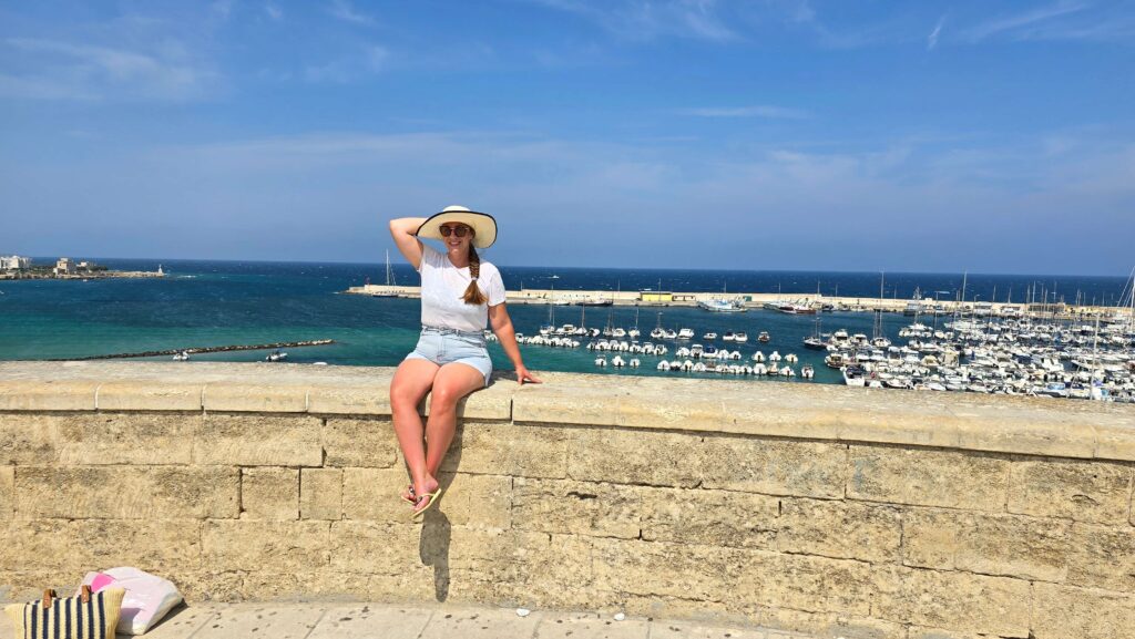 Woman in a straw hat sitting on a stone wall overlooking Otranto's harbor and the Adriatic Sea.