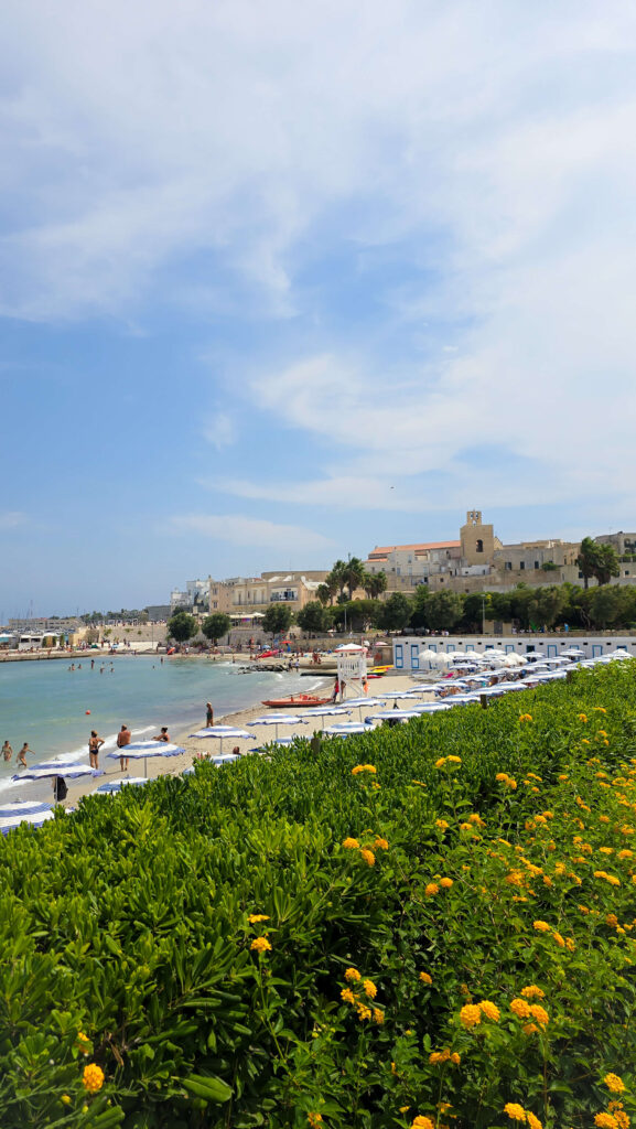 Otranto city beach with striped umbrellas, swimmers, and historic buildings in the background.