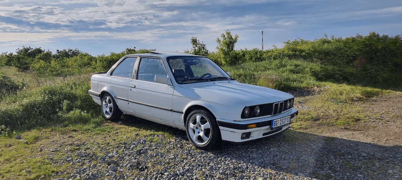 White classic BMW E30 coupe parked on a gravel roadside with green foliage under a sunny sky, ideal for European road trips