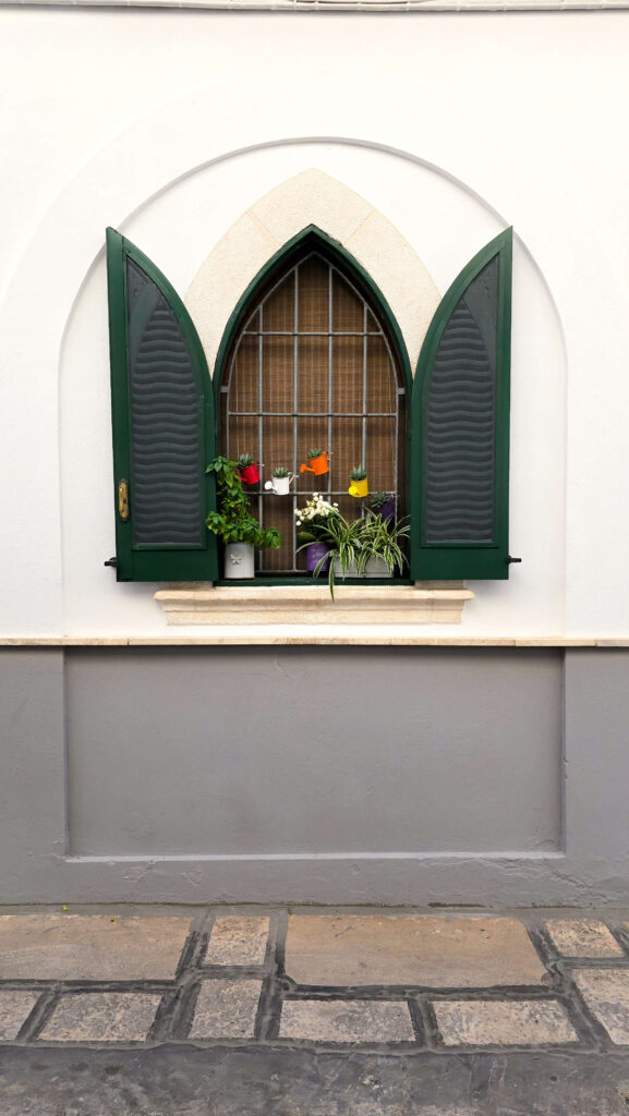 White building facade with a gothic-style window, dark green shutters, and potted plants in Ostuni.