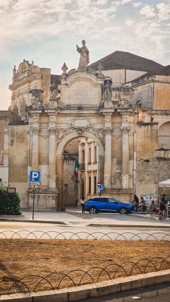 Porta Rudiae, a grand baroque city gate in Lecce, Puglia, with a blue car passing through.