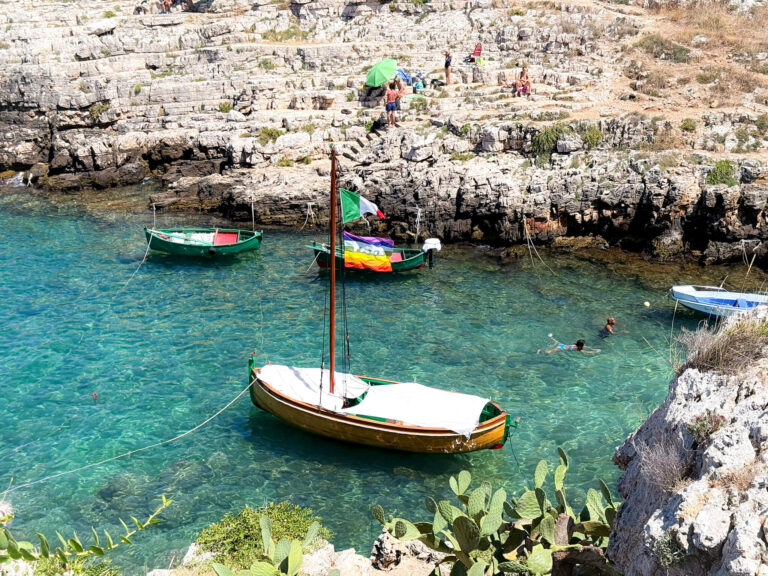 Small boats with Italian and rainbow flags in clear turquoise water at a rocky beach in Monopoli, Puglia.