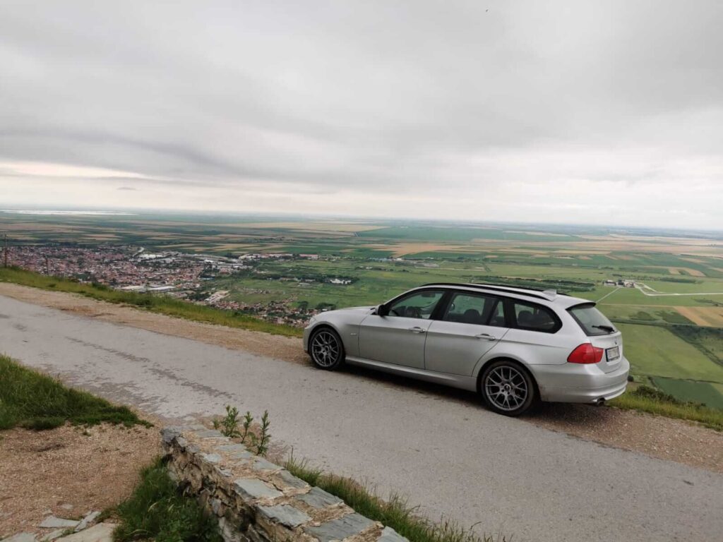 Silver car parked on a roadside overlooking a vast, green Serbian landscape and a town below