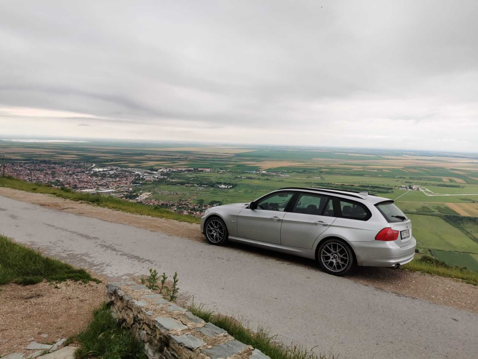 Silver car parked on a roadside overlooking a vast, green Serbian landscape and a town below. The best travel stories and European road trips are with BMW.