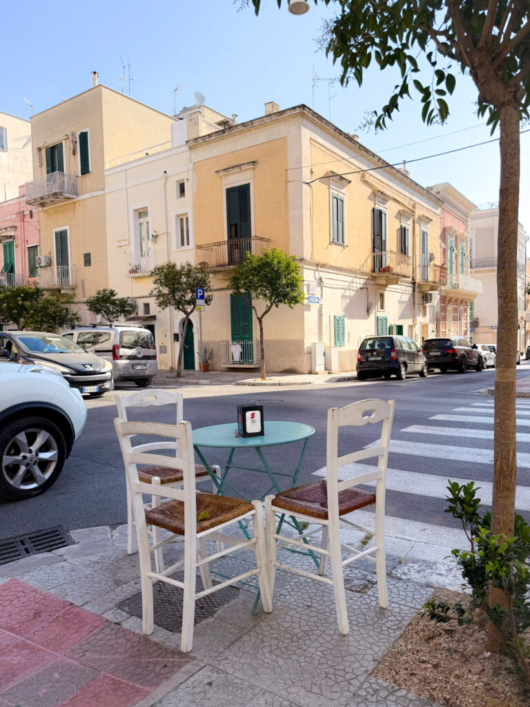 Outdoor cafe table with two white chairs on a street corner in Monopoli, Puglia, with traditional buildings.