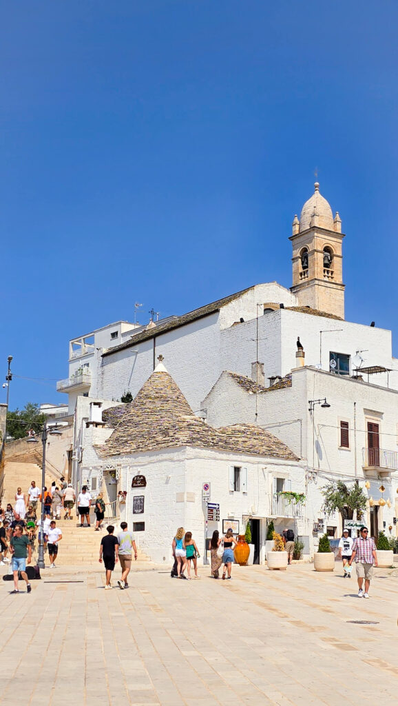 Iconic Trullo house with a conical roof in Alberobello, Puglia, with a church bell tower in the background.