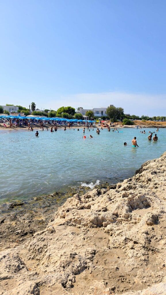 A sandy beach near Ostuni, Puglia, with a rocky foreground and people swimming in the clear blue water.