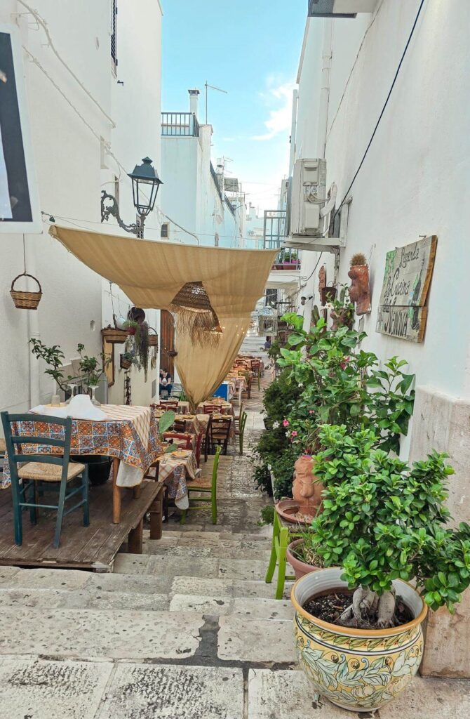 A narrow, white-walled street in Ceglie Messapica, Puglia, lined with small restaurants and lush green plants.