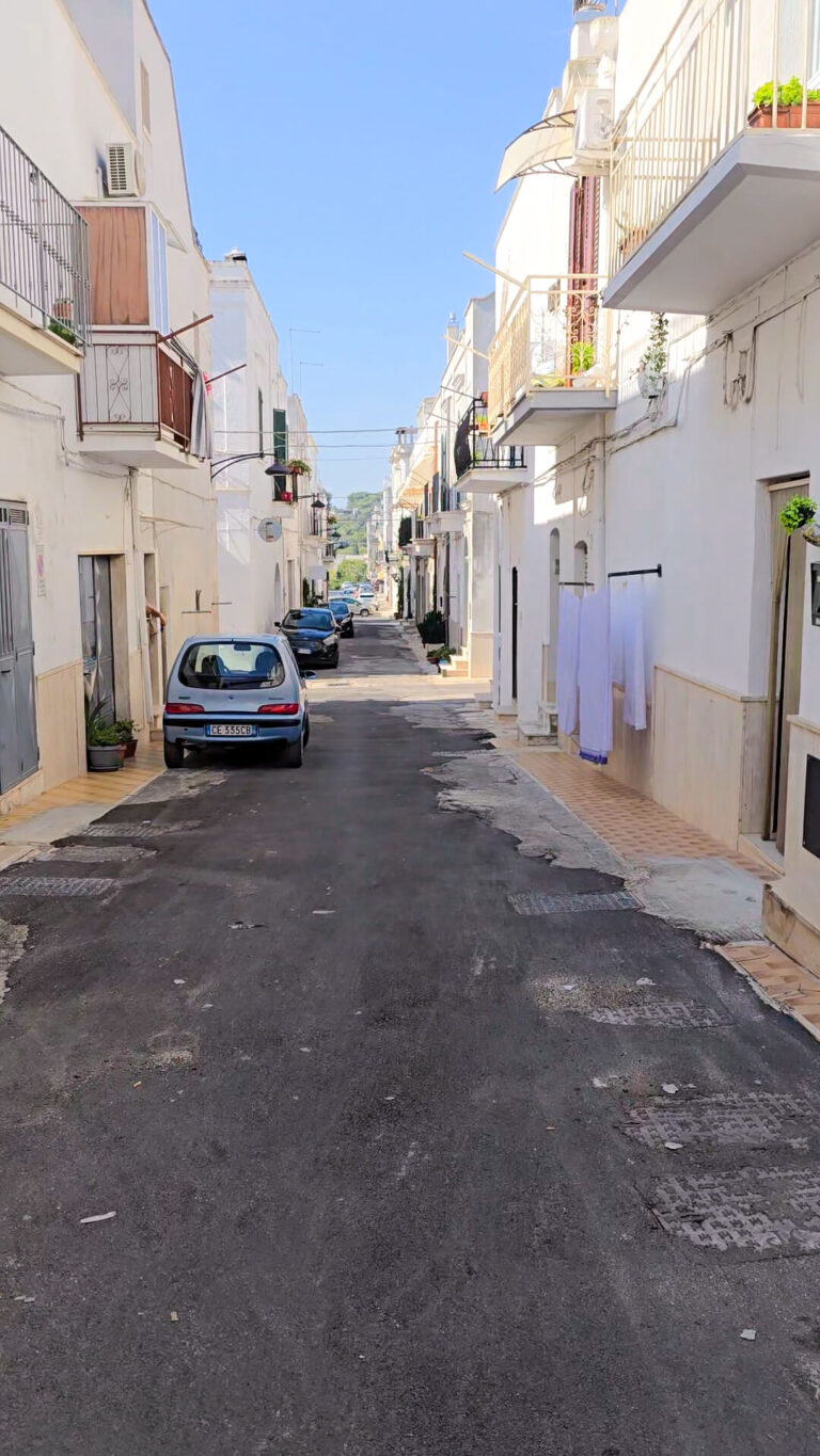 A quiet, residential street in Ceglie Messapica, Puglia, with white buildings and a parked car.