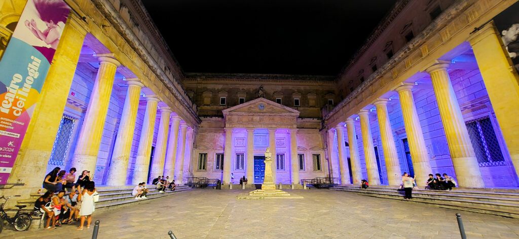 A grand, historic building with large columns and steps, illuminated with purple and yellow lights, at night. People are sitting on the steps.