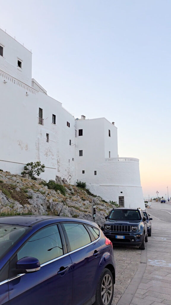 A view of the white buildings of Ostuni, Puglia, from a road with parked cars in the foreground. This is a Guide to Ostuni perfect image