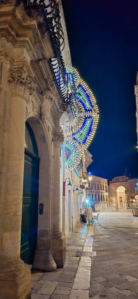 A large, ornate light decoration hangs from a balcony on a historic stone building in Lecce, Italy, at night. Presenting a guide to Lecce