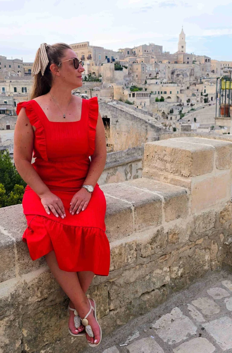 Beca from Road Sips travel blog sitting on a stone wall in Matera, Italy, overlooking historic buildings at sunset.