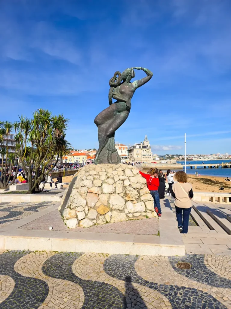 Statue by the seaside in Cascais, Portugal, with ocean views and colorful houses in the background.