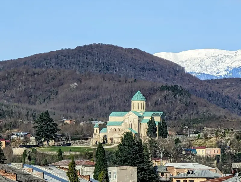 A stunning panoramic view of Bagrati Cathedral with its turquoise dome, set against the backdrop of forested hills and snow-capped Caucasus mountains, a key highlight of any Kutaisi travel guide.