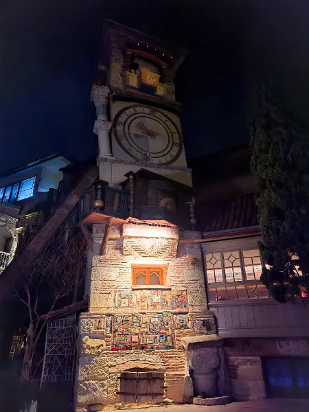 The leaning Gabriadze Clock Tower in Old Tbilisi at night with its golden clock face illuminated.