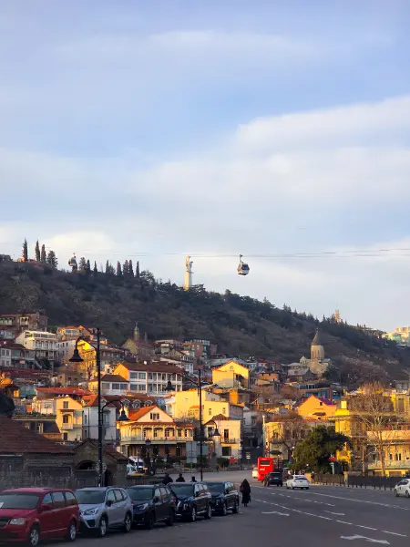 A golden-hour view of old Tbilisi used in a guide on how to get from Kutaisi to Tbilisi, showing the city’s cable car passing over traditional colorful houses and the Mother of Georgia statue on the hill.